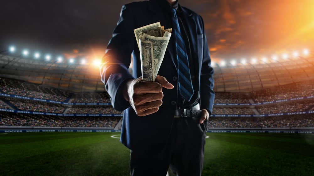 man in suit holding US dollar bills while standing in a soccer stadium